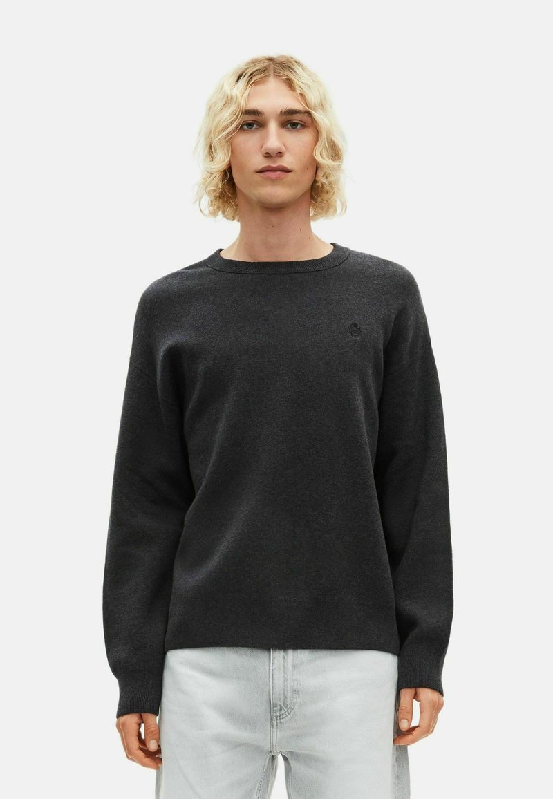 Maglione grigio scuro realizzato in materiale morbido, con scollo rotondo, maniche lunghe e un piccolo logo sul petto. Abbinato a jeans chiari.