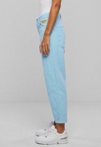 Pantaloni in velluto a coste azzurro chiaro con taglio dritto, tasche frontali e dettaglio ricamato arancione in vita, abbinati a sneakers bianche.