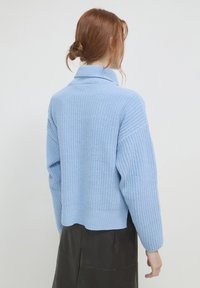 Helles blaues Strickpullover mit hohem Kragen, überschnittenen Schultern und lockerer Passform, mit Rippstruktur und einem Saum, der über der Taille endet.