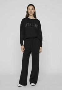 Juodas džemperis su "ATELIER" tekstu kontrastinga šviesia siūle, derinamas su laisvomis juodomis kelnėmis, pagamintomis iš minkštos medžiagos.