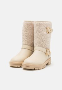 Bottines en cuir beige avec un dessus en fausse fourrure de couleur crème, dotées d'une boucle dorée en forme de cœur et d'une semelle en caoutchouc texturée.