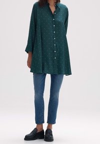 Camicia verde con bottoni e maniche lunghe, caratterizzata da un motivo sottile di piccole forme blu, abbinata a jeans skinny blu e mocassini neri.