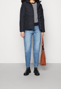 Schwarze gepolsterte Jacke mit Kapuze, gestreifter Pullover, hellblaue Jeans und schwarze Stiefeletten. Braune Tragetasche mit Griffen.