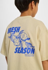 Beżowa bawełniana koszulka z niebieską grafiką piłki nożnej oraz pomarańczowym napisem „FRESH SEASON” i „hummel sportswear” na plecach.