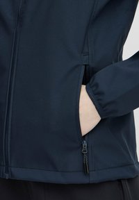 Mano nella tasca di una giacca blu scuro con zip visibile e polsino elasticizzato sulla manica, su sfondo chiaro.