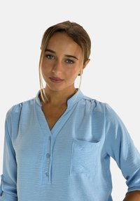 Jasnoniebieska bluzka z teksturowanej tkaniny, zapinana na guziki z przodu, z małą kieszonką na lewej piersi. Posiada kołnierz i podwinięte rękawy.