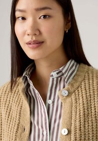 Cardigan beige lavorato a maglia con texture a coste, con una camicia a righe blu chiaro e bordeaux sotto. Bottoni bianchi mettono in risalto il cardigan.