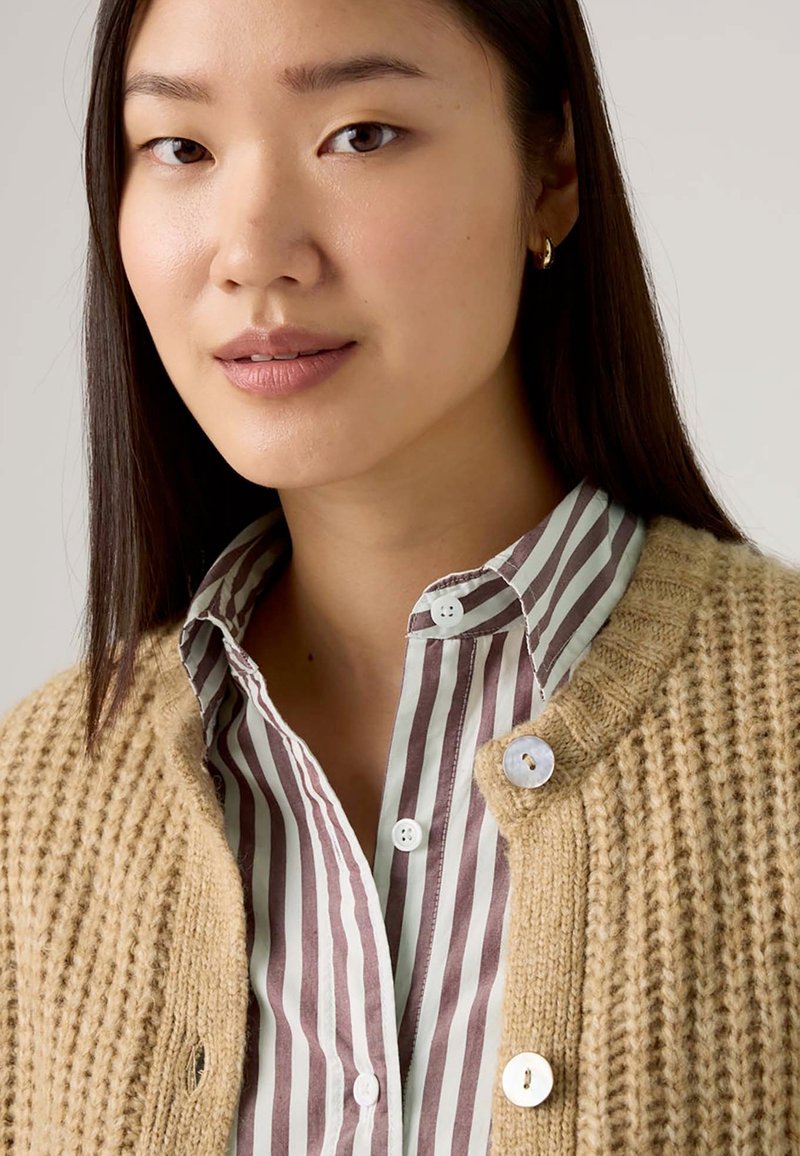 Cardigan beige lavorato a maglia con texture a coste, con una camicia a righe blu chiaro e bordeaux sotto. Bottoni bianchi mettono in risalto il cardigan.
