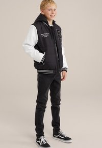 Jachetă puffer neagră cu mâneci albe, glugă și manșete cu dungi. Prezintă text brodat și un design cu logo. Este asortată cu blugi negri și adidași.