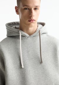 Felpa con cappuccio grigia dal texture morbido, con lacci leggeri e tasca a marsupio. Presenta una vestibilità comoda e un design minimalista.
