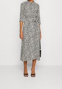 Robe florale noire et blanche avec des manches trois-quarts, taille nouée, longueur midi et une silhouette fluide. Assortie avec des talons ouverts noirs.