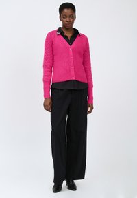 Heller pinker Strickcardigan mit strukturierten Ärmeln, V-Ausschnitt und Knöpfen. Tragen über einem schwarzen Shirt und kombiniert mit schwarzen weit geschnittenen Hosen.