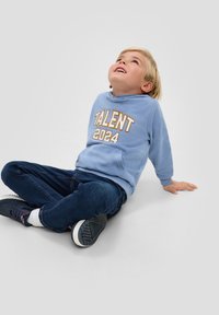Sudadera azul claro con "TALENTO 2024" impreso en un bold naranja, combinada con jeans oscuros y zapatos navy, sentado sobre un fondo gris claro.