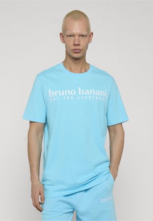 Junge Person mit kurzen blonden Haaren, die ein hellblaues "bruno banani" T-Shirt und passende Shorts trägt, mit den Händen in den Taschen vor schlichtem Hintergrund.