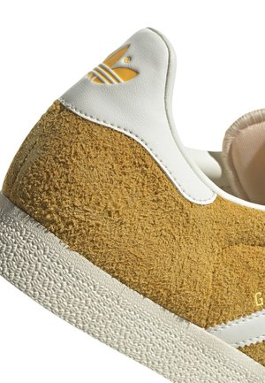 Gros plan sur le talon d'une basket en daim beige avec un col en cuir blanc et le logo Adidas jaune en relief, montrant une semelle blanche texturée.