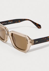 Sunglasses s priehľadnými béžovými štvorecovými rámami a ramienkami s korytnačkovým vzorom ležiace na bielej ploche.