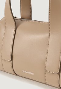 Beige læderhåndtaske med dobbelte håndtag, lynlåslukning og præget Calvin Klein-logo. Glat tekstur med kontrastsyninger.