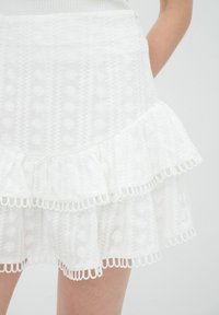 Jupe blanche avec un design à volants superposés, ornée de broderies florales et d'une finition en dentelle festonnée le long de l'ourlet. Tissu léger et texturé.