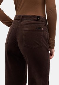 Braune Cordhose mit Gesäßtaschen, darüber ein schwarzes Etikett auf der rechten Tasche und einem klassischen, geraden Schnitt.