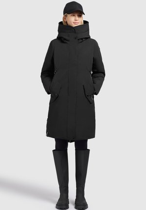 Parka nera isolata con colletto alto, tasche laterali e chiusura con bottoni. Indossata con leggings neri e stivali di gomma al ginocchio.