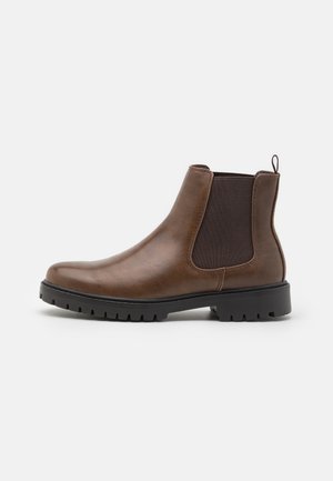 Botte Chelsea en cuir marron avec panneaux latéraux élastiques noirs, languette à l’arrière et semelle épaisse noire à crampons sur fond blanc.