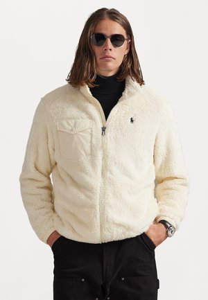 Fleece jacket in cream with a zip front, high collar, and chest pocket. Features a black logo on the left side. Worn over a black turtleneck.