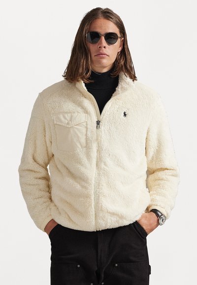 Fleecejacke in Creme mit Reißverschluss, hohem Kragen und Brusttasche. Verfügt über ein schwarzes Logo auf der linken Seite. Getragen über einem schwarzen Rollkragenpullover.