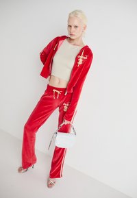 Punainen samettinen urheilasu kultaisilla yksityiskohdilla, vetoketjullinen huppari ja siihen sopivat housut, joissa on sivuraidat. Pieni valkoinen käsilaukku ja hopeiset sandaalit.