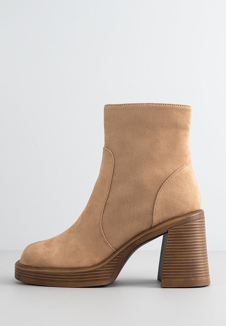 Bottine en daim beige à bout rond, avec des panneaux élastiques sur les côtés et un talon en bois nervuré. Texture lisse et design épuré.