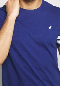 Modrá bavlněná tričko s kulatým výstřihem, bílým logem na hrudi a pruhovaným bílým detaily na rukávech. Hladká textilie.
