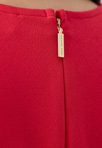 Červená textúrovaná tkanina s vertikálnym švom a zlatým zipsom. Na ťaháku zipsu je vyryté meno "MICHAEL KORS."