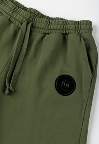 Olejkowe dresy z elastycznym paskiem w talii, sznurkiem do regulacji oraz okrągłą czarną naszywką z wzorem i logo na lewym udzie.