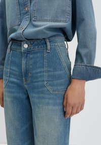 Jean en denim bleu clair taille haute avec un design à cinq poches, des coutures visibles et une fermeture à bouton, associé à une chemise en denim assortie.