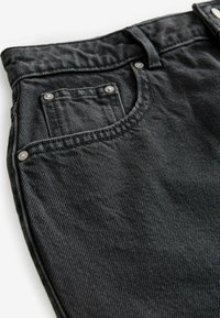 Jeans de mezclilla negra con un acabado texturizado, que cuentan con un bolsillo frontal con remaches metálicos y una cremallera. La vista en primer plano enfatiza los detalles de la tela y la costura.