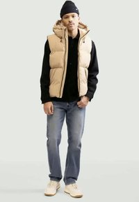 Beige puffer vest with a hood over black shirt, paired with blue jeans and light sneakers. Simple design, warm material, and contrasting textures.