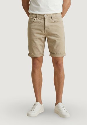 Uomo con pantaloncini beige arrotolati, sneakers bianche e t-shirt bianca, in piedi contro uno sfondo chiaro uniforme.