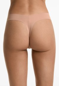 Nude tanga ondergoed gemaakt van glad, rekbaar materiaal met een minimalistisch ontwerp, voorzien van een naadloze tailleband en zonder zichtbare lijnen.