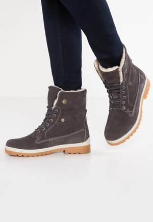 Szare zimowe botki ze skóry zamszowej na sznurowadła, z białym futrzanym wykończeniem i jasnobrązowymi gumowymi podeszwami, noszone z ciemnymi dżinsami, jedna stopa uniesiona w półkroku na białym tle.