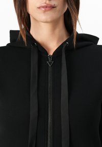 Zwarte hoodie met een ritssluiting aan de voorkant, contrasterende trekkoord en een gladde textuur. Opvallend driehoekig ritssleepdetail.