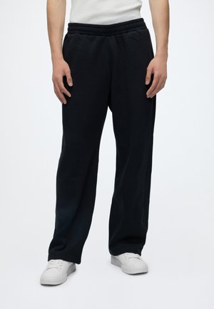 Personne portant un pantalon de jogging noir ample, des baskets blanches et une chemise blanche, debout devant un fond clair uni.