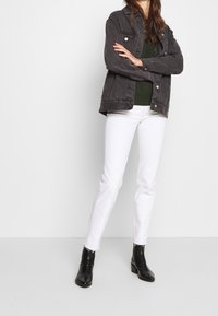 Mujer de pie con los brazos cruzados, vestida con una chaqueta de mezclilla oscura, un suéter verde, jeans blancos y botas negras hasta el tobillo, sobre un fondo blanco.