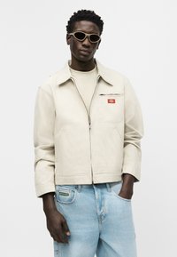 Homme portant une veste zippée en velours côtelé crème, une chemise beige, un jean bleu clair et des lunettes de soleil ovales beige, debout avec une main dans la poche.