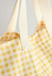 Sac cabas en tissu à carreaux jaunes et blancs avec de larges anses texturées couleur blanc cassé sur un fond uni.