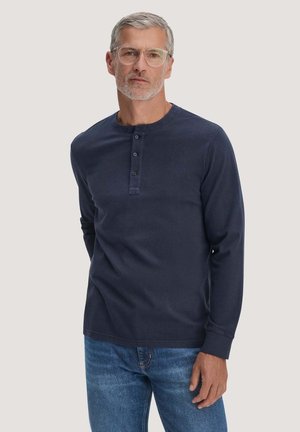 Mittelalter Mann mit grauen Haaren und Brille, der ein marineblaues Henley-Shirt mit langen Ärmeln und blaue Jeans vor einem einfarbigen Hintergrund trägt.