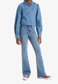 Camisa de mezclilla azul con mangas abullonadas y dos bolsillos en el pecho, combinada con jeans acampanados en azul claro. Completado con zapatillas de lona rojas y blancas.