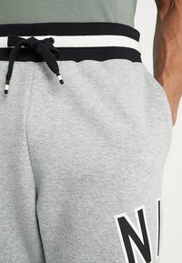 Detailní záběr na osobu, která má na sobě světle šedé tepláky s černobílým stahovacím pasem a částečným černým písmenem "N" na nohavici.