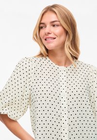 Νέα γυναίκα με ξανθά μαλλιά που φοράει λευκό πουκάμισο με μαύρες πουά και φουσκωτά κοντά μανίκια, χαμογελά και κοιτάζει στο πλάι.