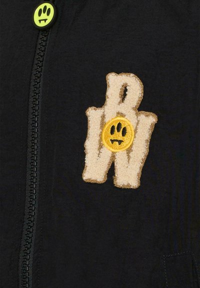 Giacca nera con zip caratterizzata da un logo ricamato beige testurizzato con un accento di faccina sorridente gialla. Bottone verde vicino al colletto.