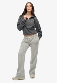 Pull en gris avec col zippé, arborant le texte "Superdry Athletics". Pantalons de survêtement gris clair avec logo, coupe décontractée et taille élastique avec cordon de serrage.