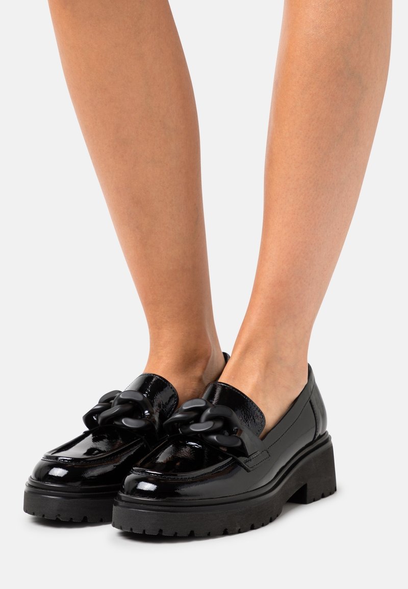 Mocassins en cuir noir brillant avec semelles plateformes épaisses et détail de chaîne surdimensionnée, portés pieds nus sur fond blanc.
